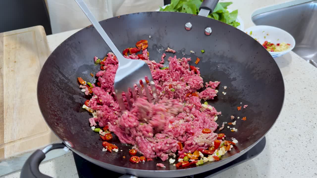 Raw minced meat is stirred with chopped chili and vegetables in a wok under bright kitchen lighting, highlighting the initial stage of spicy stir-frying