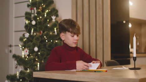 Junge-Im-Roten-Rollkragenpullover-Faltet-Einen-Wunschbrief-Und-Steckt-Ihn-In-Einen-Umschlag,-Der-An-Einem-Tisch-In-Einem-Mit-Einem-Weihnachtsbaum-Geschmückten-Raum-Sitzt