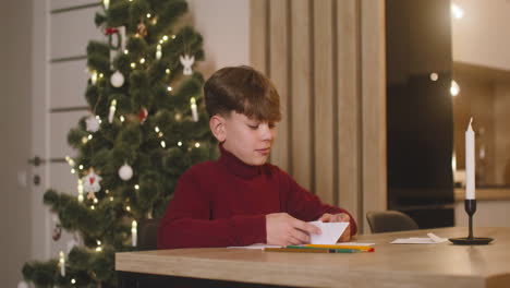 boy in red turtleneck folding a letter of wishes and inserting it in a evelope sitting at a table in a room decorated with a christmas tree