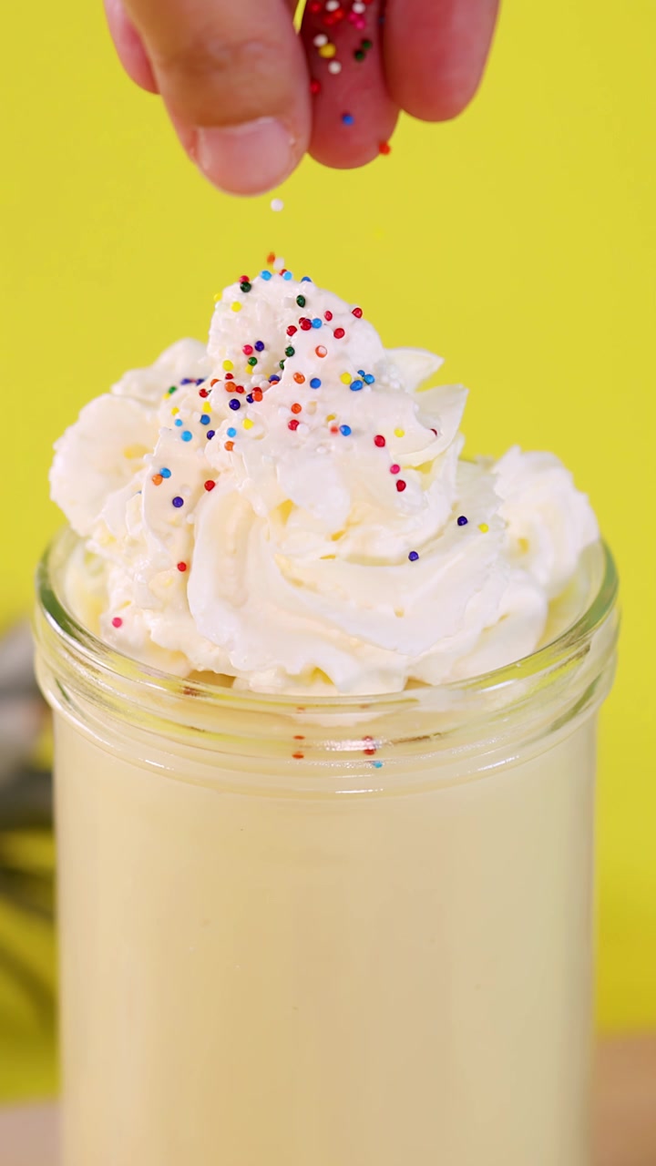 Primo piano di una mano che sparge caramelle colorate su un frullato all'ananas con panna montata, su uno sfondo giallo brillante.