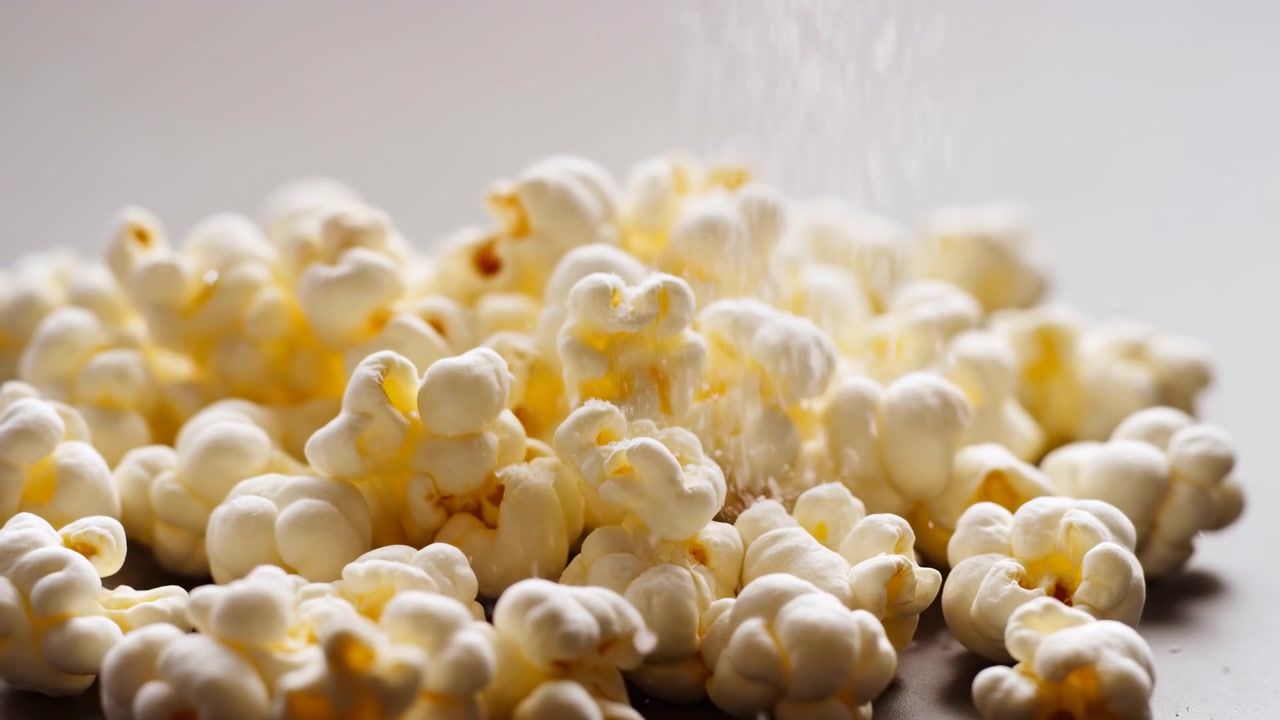 Vista ravvicinata dal basso di popcorn ammucchiati su una superficie piana, con particelle di condimento visibili che cadono dall'alto, morbidi riflessi dall'alto, texture cremosa, dettagli nitidi e caldi.