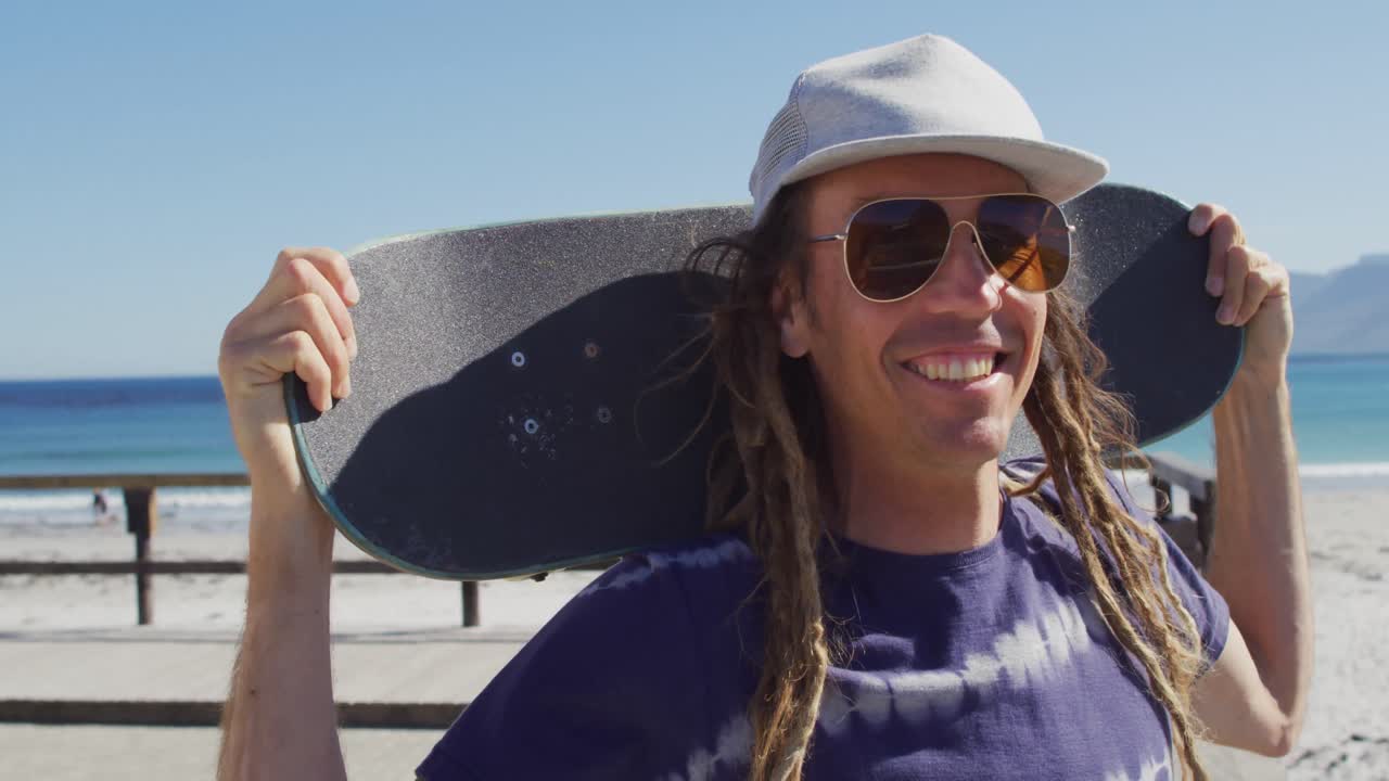 retrato de video de un hombre caucásico sonriente con dreadlocks sosteniendo un skateboard en un paseo marítimo soleado