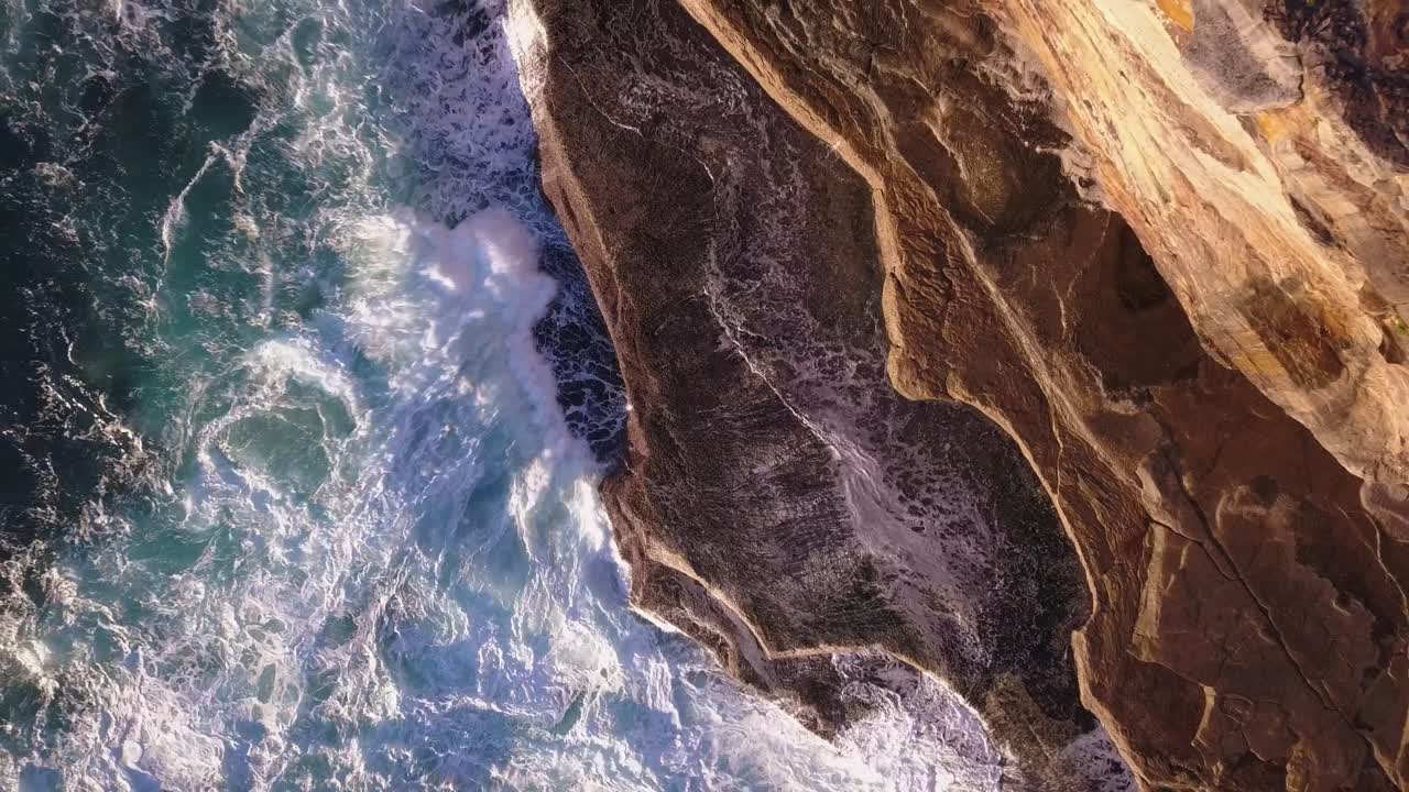 Potenti onde dell'oceano che si infrangono su rocce e scogliere, ripresa aerea dall'alto verso il basso con movimento ascendente che rivela la scena