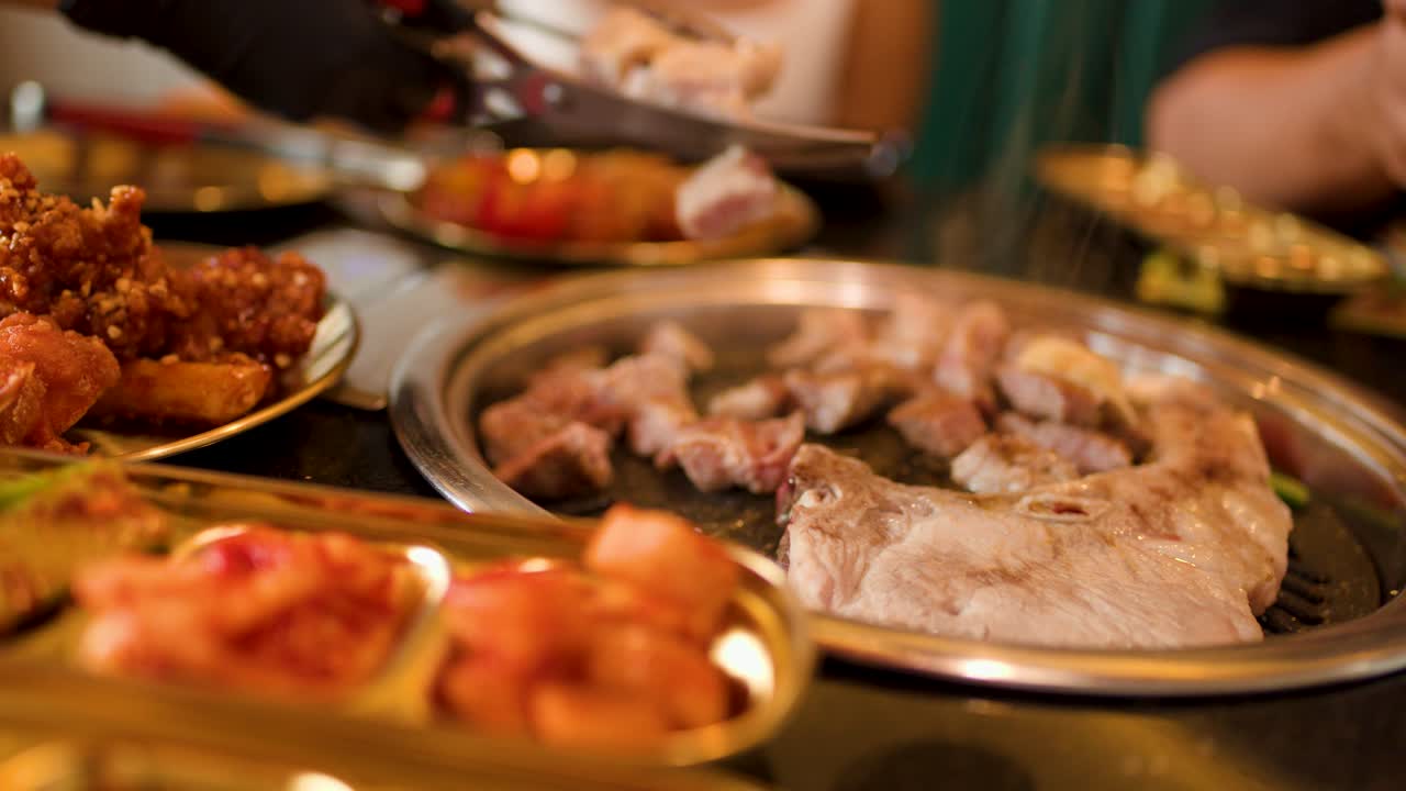 Close-up of assorted meats cooking on grill pan, hands using tongs, warm indoor lighting