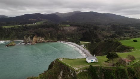 aerial backwards of a small hermitage surrounded by nature, ermita de la regalina, situated on the steep cliffs of the cantabrian sea in asturias, spain