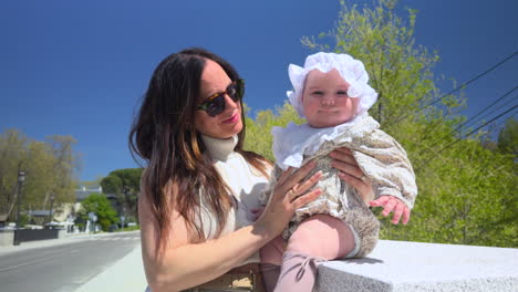 young mother lovingly holds her smiling baby daughter during a sunny spring day, standing near a quiet street with trees and blue sky in the background