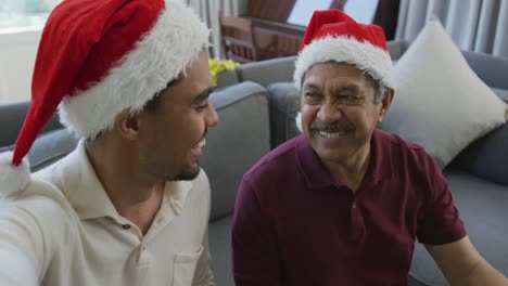 Retrato-De-Un-Hombre-Alegre-Mostrando-Un-Regalo-Mientras-Está-Sentado-Junto-A-Un-Hijo-Birracial-Haciendo-Una-Videollamada-Navideña