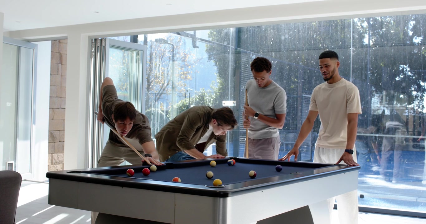 Diverse male friends watching left player lining up and taking shot, playing pool in rec room