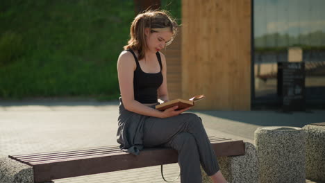 dama sentada al aire libre con las piernas cruzadas, leyendo un libro mientras el viento sopla a través de su cabello y las páginas del libro, la luz del sol se refleja en ella, con un fondo borroso con una escalera