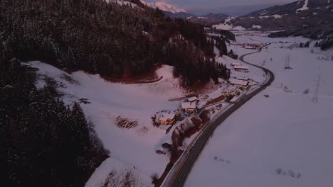 Toma-Aérea-De-Un-Pueblo-De-Montaña-Cubierto-De-Nieve,-Camino-Sinuoso-A-Través-Del-Valle-Durante-La-Puesta-De-Sol