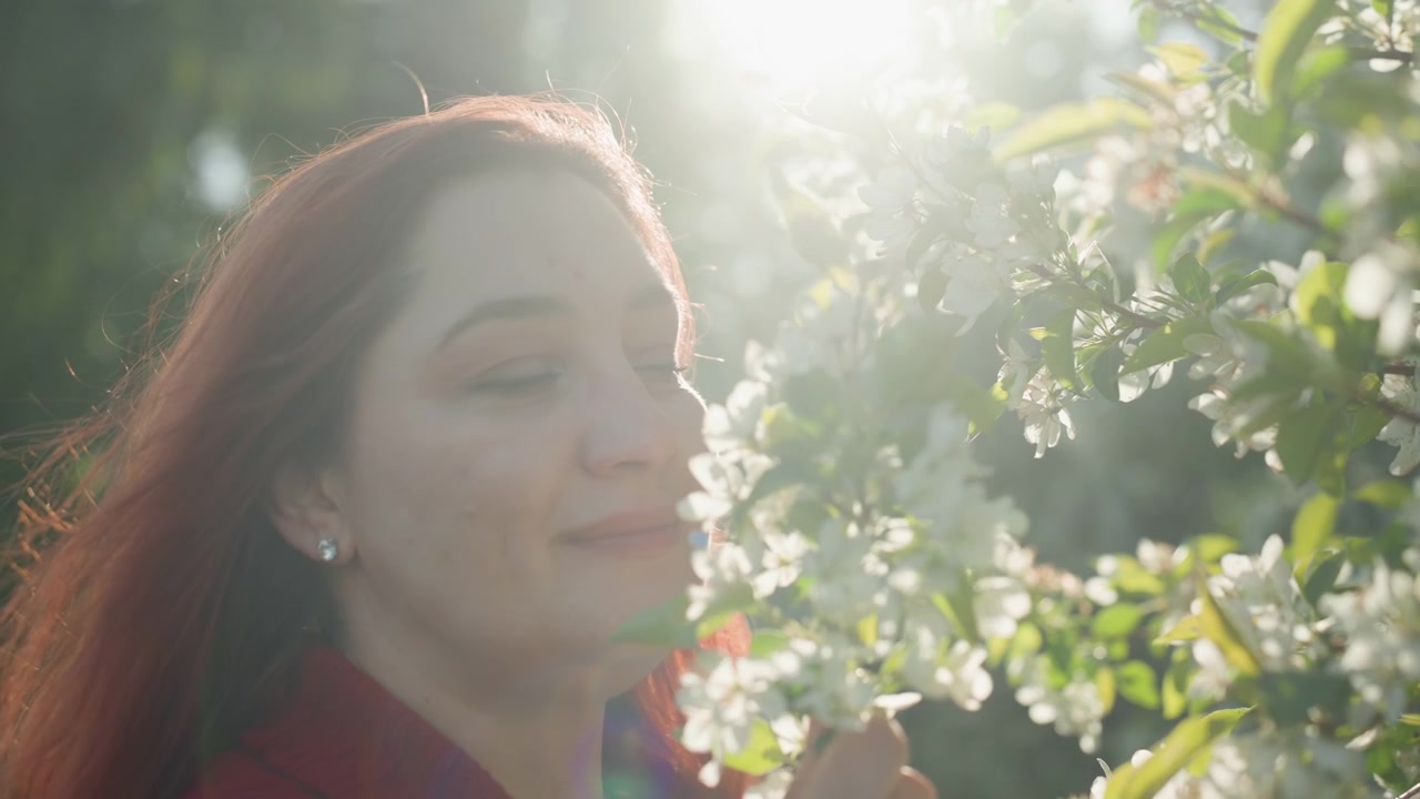 Primo piano di donna con lunghi capelli rossi che indossa orecchini e annusa fiori bianchi alla luce del sole. La scena presenta un'illuminazione soffusa, uno sfondo di fogliame verde e il soggetto che tiene i fiori vicino al viso.
