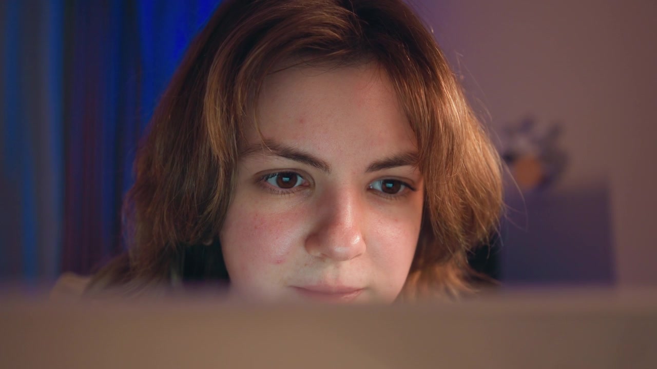 Close view of young woman leaning toward foreground screen with narrowed eyes and slight furrowed brow, showing concentration, engagement, warm frontal light, cool blue curtain backdrop and quiet