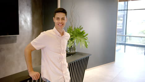 smiling man standing in modern office lobby, leaning on table with plant, copy space