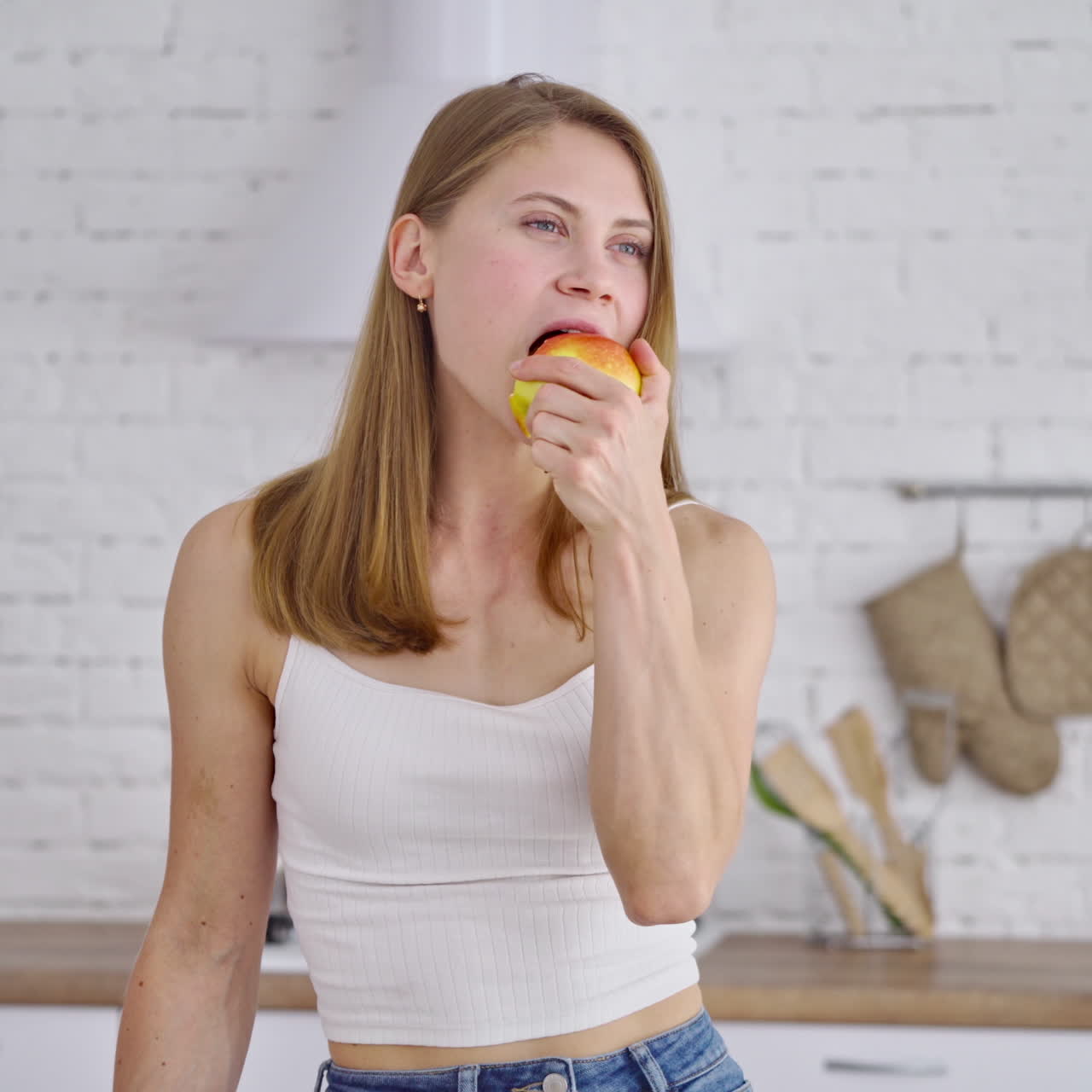 Beautiful woman eating tasty apple indoors. Young female with sporty body standing in the modern kitchen and eats fresh fruit. Portrait of a lovely girl looking aside.