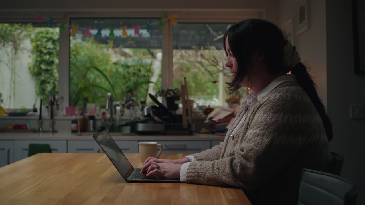 A middle-aged woman smiles while working on her laptop in a cozy kitchen, using an AI chatbot for family budgeting. Warm, homely atmosphere with a relaxed mood.