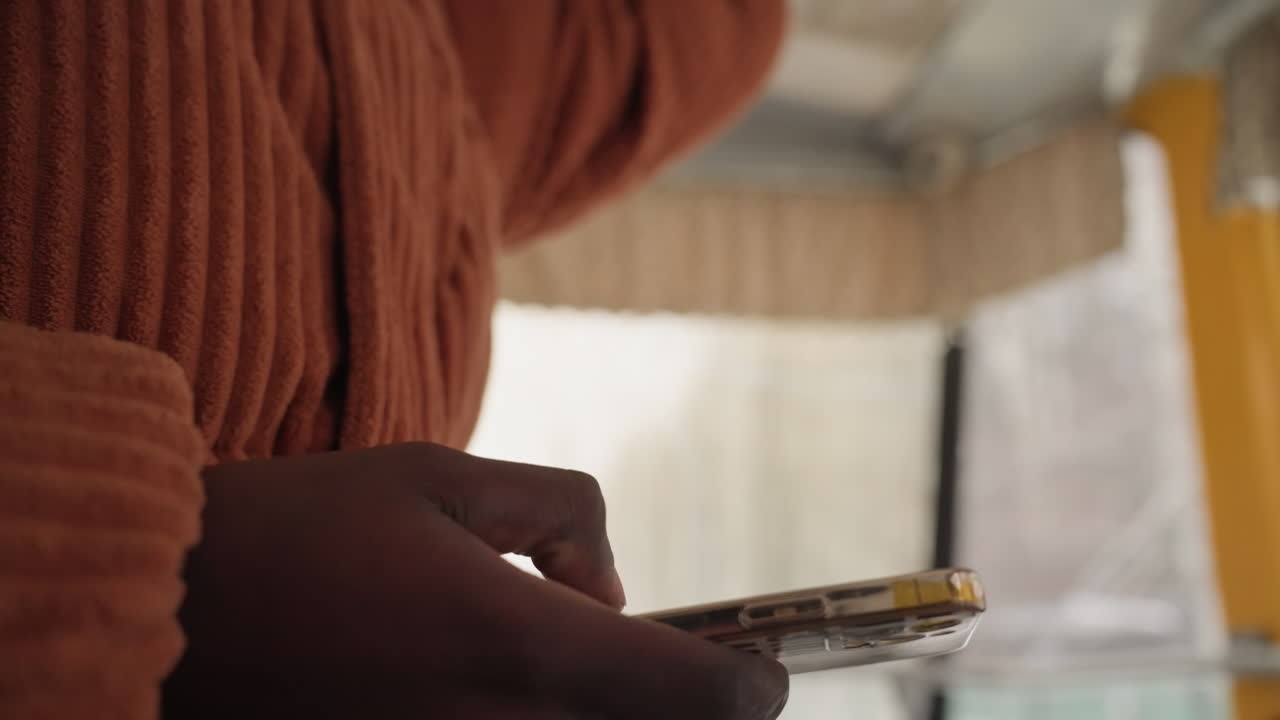 Man hands holding phone closeup inside bus, orange knit sweater, tapping and scrolling through messages, checking ticket and map, soft natural light through window, candid intimate frame showing