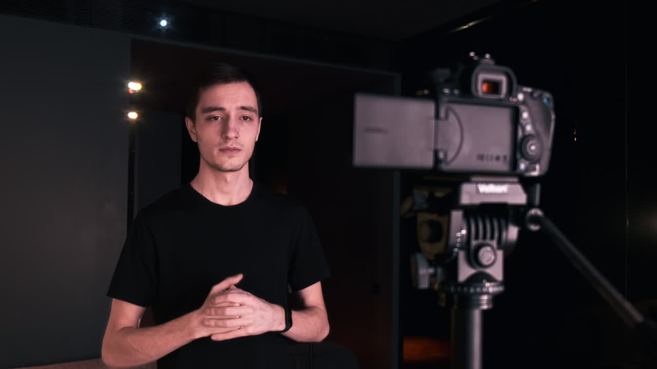 Un giovane creator di contenuti parla e gesticola mentre si riprende usando una fotocamera su un treppiede durante la registrazione di un vlog. Lavora da casa