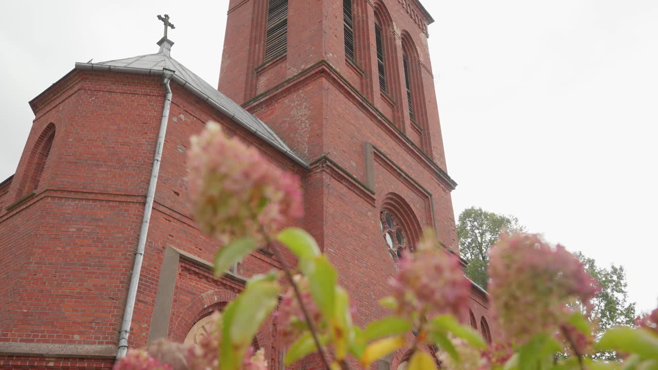 Immagine di una storica chiesa in mattoni rossi con un primo piano fiorito. La composizione verticale contrasta magnificamente i dettagli architettonici con morbidi elementi floreali