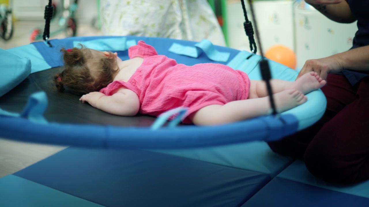 Baby girl wearing a pink romper lying on a mat, receiving gentle physiotherapy from a healthcare professional for motor skills