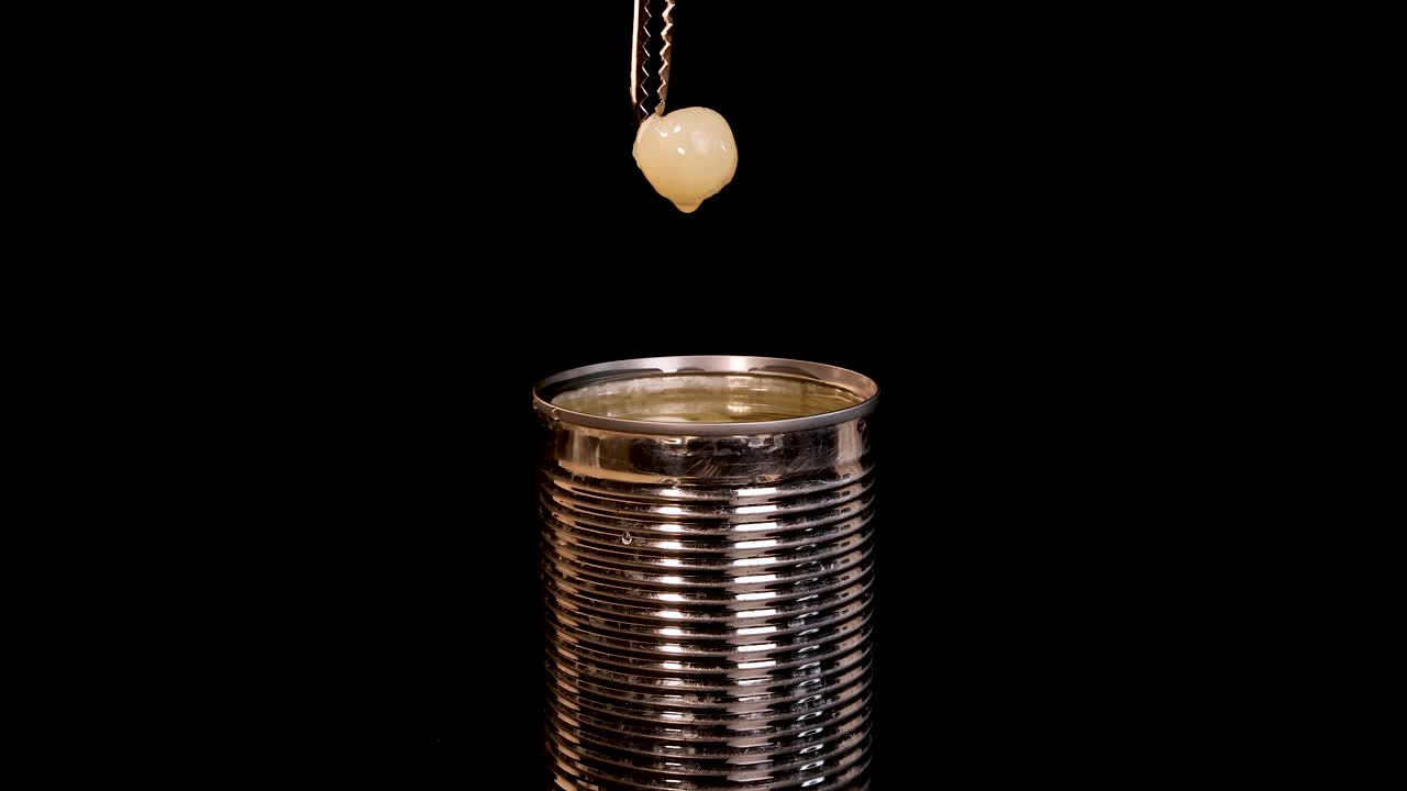 A hand uses metal tongs to lift a peeled longan fruit from an open canned food container against a black background, with even studio lighting and static camera