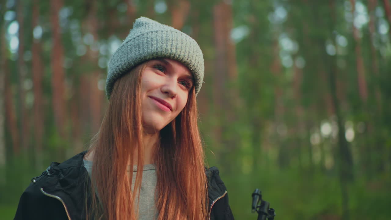 Close up of woman wearing gray knit hat and black jacket standing in forest, smiling gently while holding nozzle, glancing toward someone in background with warm, playful expression