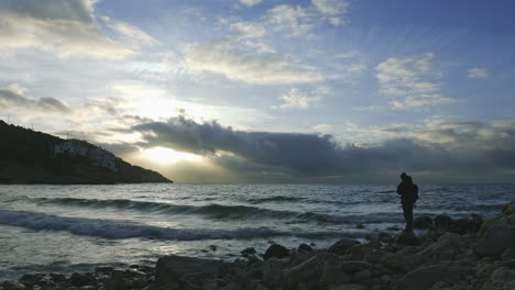 morning time lapse near rocky seaside, mans silhouette walks across, clouds moving fast sun rays breaking