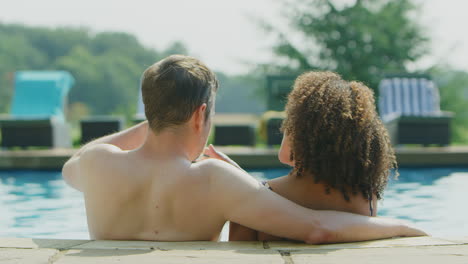 rear view of loving couple relaxing in swimming pool on vacation celebrating drinking champagne