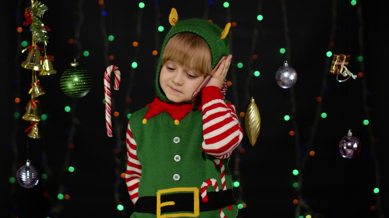 Holčička v kostýmu vánočního elfa Santa Clause pomocníka telefonuje s gratulací na mobilním telefonu
