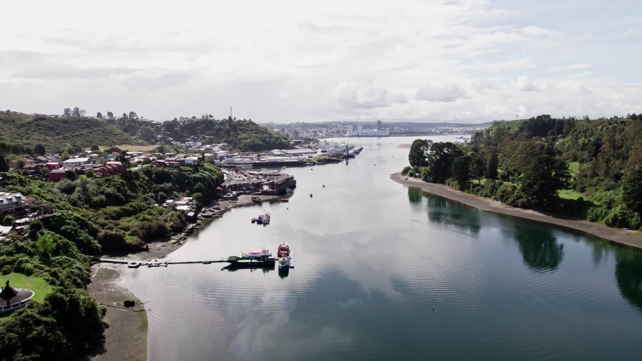 Forested, tranquil waters of the Reloncaví Sound (Chilean Fjords gateway), with a small coastal village in the foreground and the Puerto Montt city skyline visible in the distance