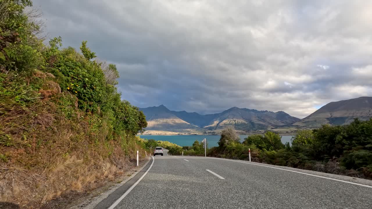 Vehicle travels winding road with lake and mountains, daylight, partly cloudy, forward-facing dashcam perspective