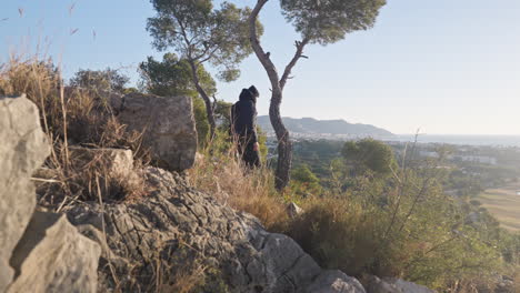 Man-climbs-down-rocky-slope,-then-stops-admires-beautiful-view-far-way-of-land,-mountains,-city-and-sea