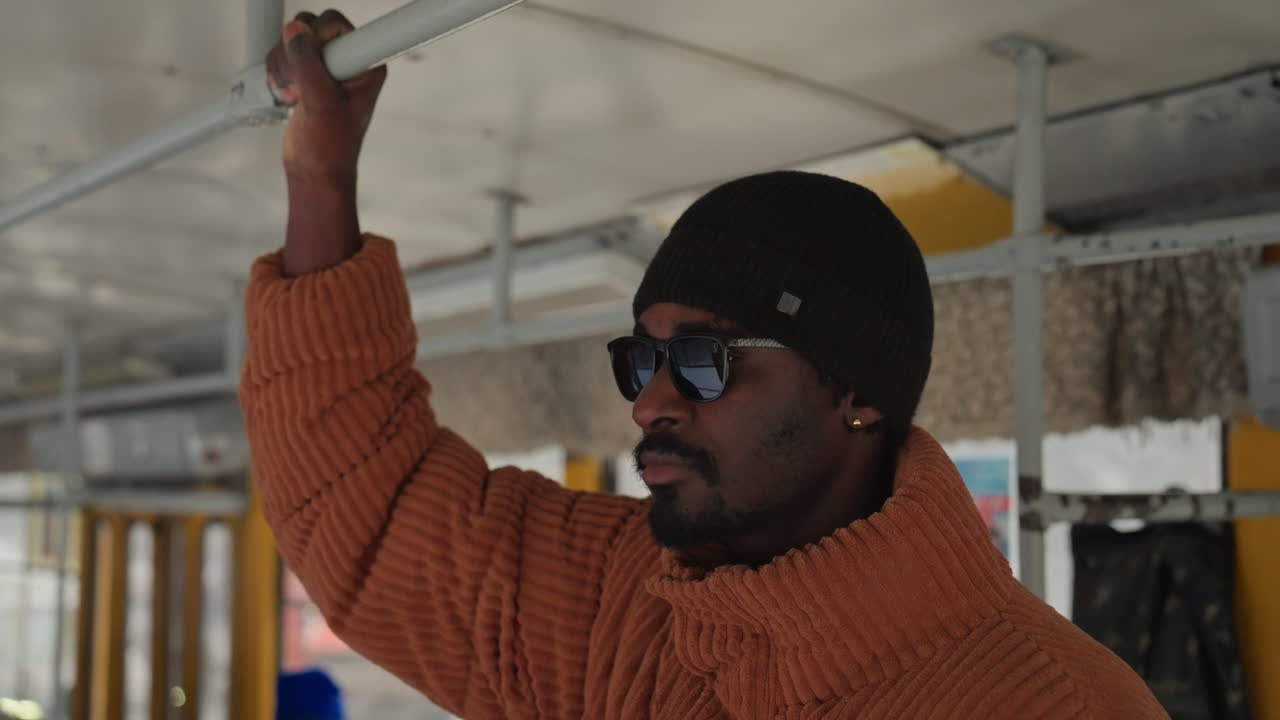 Uomo afroamericano che tiene un palo sull'autobus, berretto invernale e occhiali da sole, trama del maglione color ruggine, presa sul maniglione superiore, luce diurna attenuata attraverso i finestrini, pendolari sfocati sullo sfondo, espressione contemplativa, ambiente urbano.