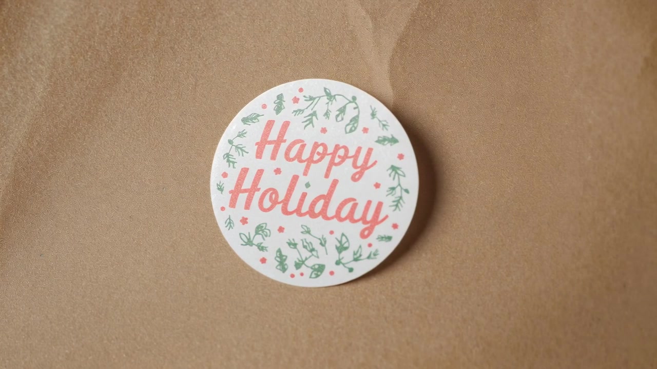 Zooming camera bringing round white sticker closer on brown kraft-paper, highlighting Happy Holiday