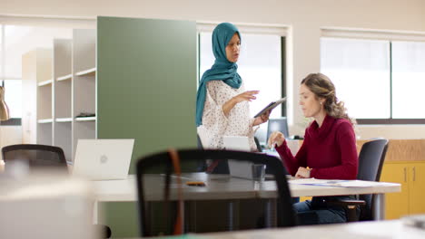 diverse female creative colleagues in discussion using laptop and tablet in office, slow motion