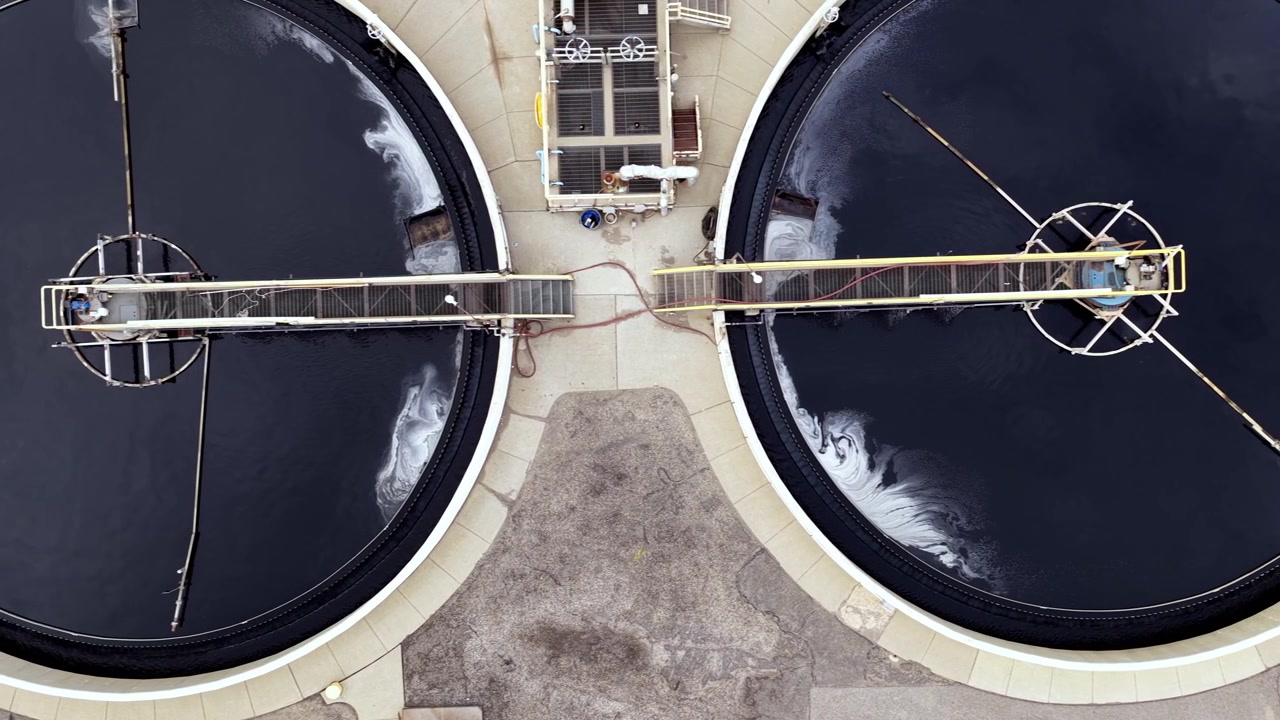Aerial footage slides over large industrial water reclamation settling tanks at a wastewater treatment plant, showcasing geometric design, infrastructure, and reflective surfaces at daytime