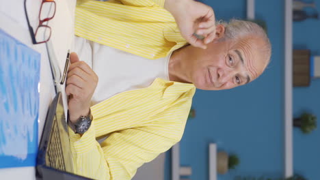 Vertical-video-of-Home-office-worker-old-man-scratches-his-ears-looking-at-camera.