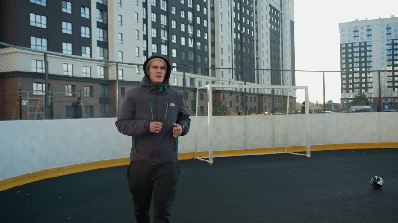 молодой человек бегает на городской спортивной арене с футбольным мячом и головным столбом на заднем плане, окруженный высотными офисными зданиями