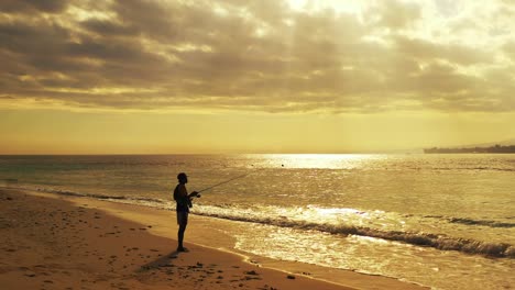 Sun-rays-showing-through-the-dramatic-clouds-above-the-sandy-beach-with-lonely-fisherman-fishing-in-the-shallow-water