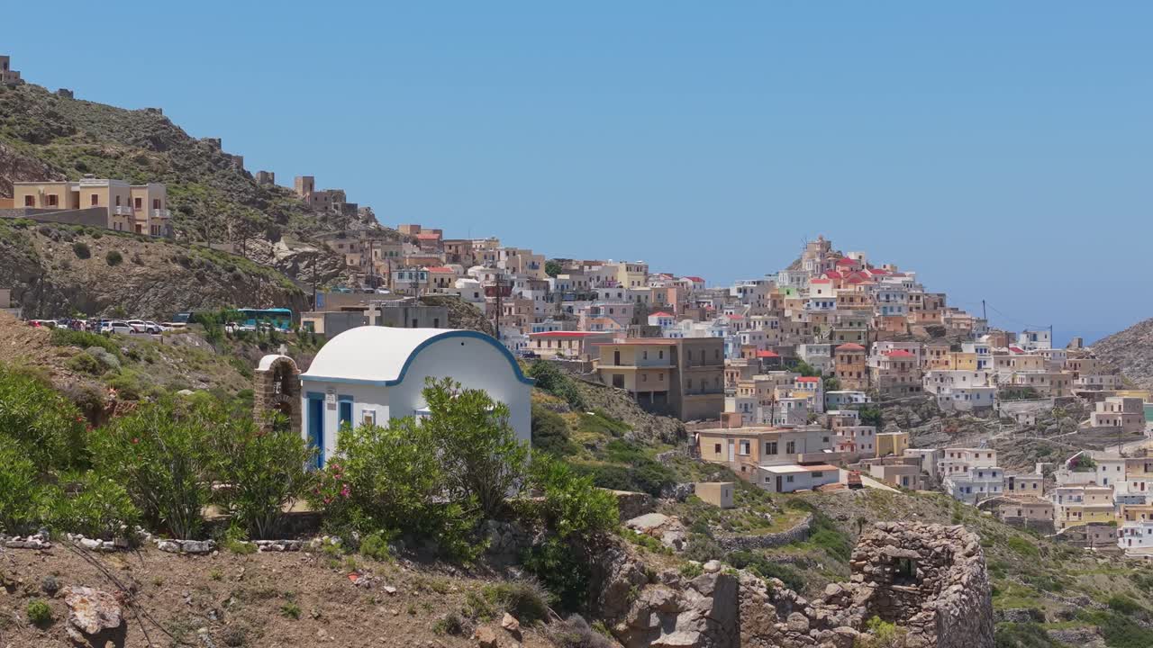 Drone orbits a whitewashed chapel in the foreground, gradually revealing the colorful traditional village of Olympos on Karpathos in the scenic hillside background