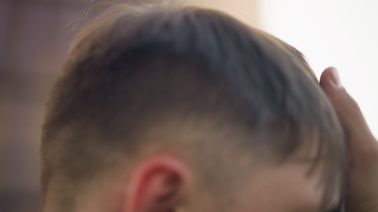 코카서스 남성이 클로즈업된 채로 왕관과 뒷머리를 만지고 있고, 은은한 창문 빛으로 내부가 밝게 빛나며, 사려 깊은 분위기, 깔끔한 페이드 컷 헤어스타일과 내성적인 제스처, 미묘한 질감과 따뜻한 색조가 돋보입니다.