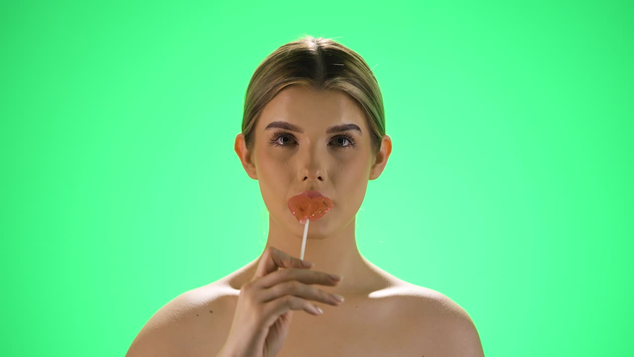 disparo medio de una mujer joven y bonita sosteniendo una paleta deliciosa en forma de corazón en la cámara antes de lamerla y disfrutar de la dulzura frente al fondo verde en cámara lenta