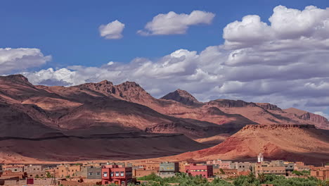 time lapse shot of flying clouds over village in morocco and red mountains in background