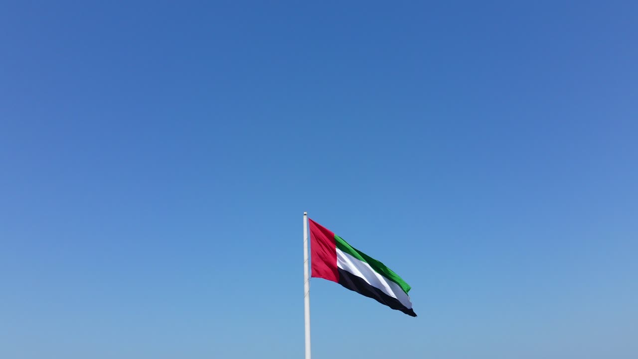 vista aérea de la bandera de los emiratos árabes unidos levantándose en un hermoso día soleado, cielo azul en el fondo, el símbolo nacional de los emiratos árabes unidos