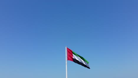 vista aérea de la bandera de los emiratos árabes unidos levantándose en un hermoso día soleado, cielo azul en el fondo, el símbolo nacional de los emiratos árabes unidos