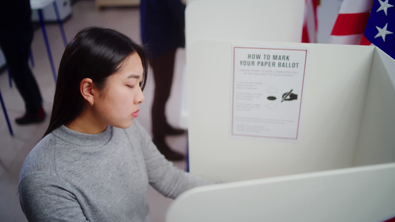 Asian Female Voter Looking at Camera Standing in Voting Booth Dolly Shot of Asian Female Voter Choosing Presidential Candidate to Vote for in Voting Booth at Polling Station then Looking at Camera us Citizen during National Election Day in the United States