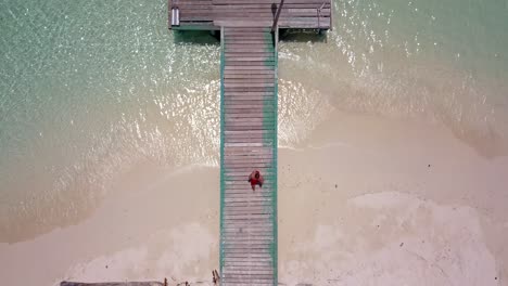 Perspectiva-Aérea-Directamente-Hacia-Abajo-En-Un-Muelle-De-Madera-En-Tailandia,-Luego-Rastreando-Al-Hombre-Mientras-Corre-Y-Se-Sumerge-En-El-Agua-Verde-Clara-Del-Océano,-Mira-Hacia-Abajo,-Sigue,-Rastrea,-4k