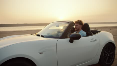 un hombre barbudo con cabello rizado en una camisa azul está conduciendo con su novia en un coche descapotable blanco en la playa contra el cielo amarillo