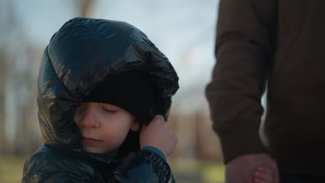 a close-up of father and son holding hands while walking outdoors, the child, dressed in a shiny black jacket and a black beanie, is seen adjusting his head warmer