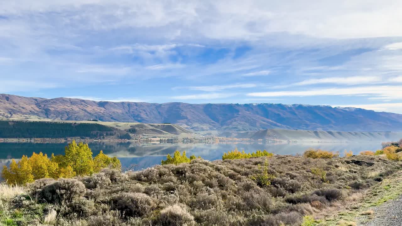 A serene drive through Queenstown, New Zealand, showcasing vibrant autumn foliage, a tranquil lake, and majestic mountains under a clear sky