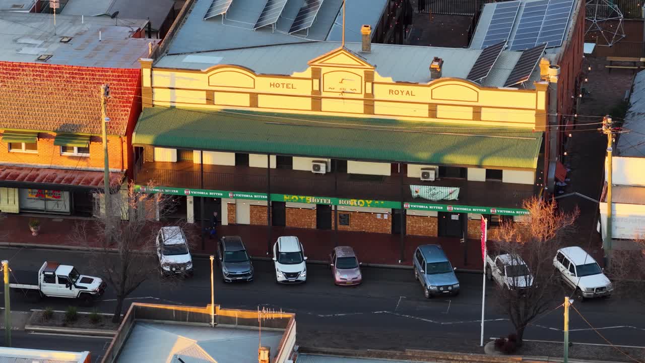 Aerial camera slowly pans across a heritage building and parked cars in Coonabarabran, Australia, during golden hour with warm sunset lighting